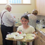 Girl carrying tray of washing up with older people in background